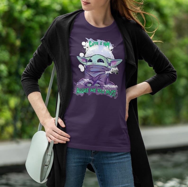 May include: A purple t-shirt featuring a graphic of a cartoon character with large ears, a green face, and a purple cloak. The text "CUTE I AM" is above the character, and "ADORE ME YOU MUST" is below. The design is set against a white cloud-like background.