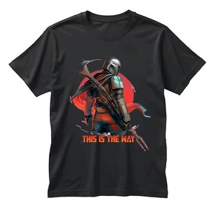 Op de afbeelding: Zwart T-shirt met een afbeelding van een Mandalorian-personage tegen een rode zon. Het personage houdt een wapen vast. De tekst "THIS IS THE WAY" is in oranje en wit gedrukt.
