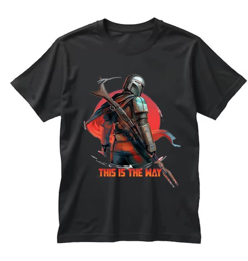 Op de afbeelding: Zwart T-shirt met een afbeelding van een Mandalorian-personage tegen een rode zon. Het personage houdt een wapen vast. De tekst "THIS IS THE WAY" is in oranje en wit gedrukt.