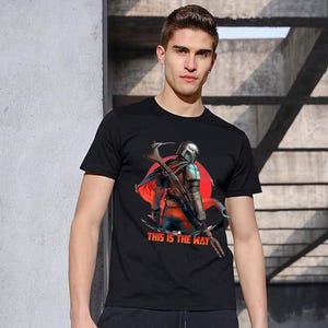 Op de afbeelding: Zwart t-shirt met een afbeelding van een Mandalorian-personage, een rode zon en de tekst "THIS IS THE WAY". Het ontwerp toont een gedetailleerde illustratie van het personage met een wapen. Het t-shirt is gemaakt van zacht materiaal.