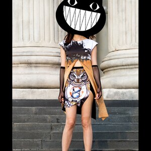 Peut inclure: Une femme portant un t-shirt graphique noir et blanc avec un masque de chat noir arborant un large sourire blanc. Elle porte une jupe marron et noire avec un motif de hibou et des gants en r&eacute;sille noirs. Elle est debout sur un escalier.