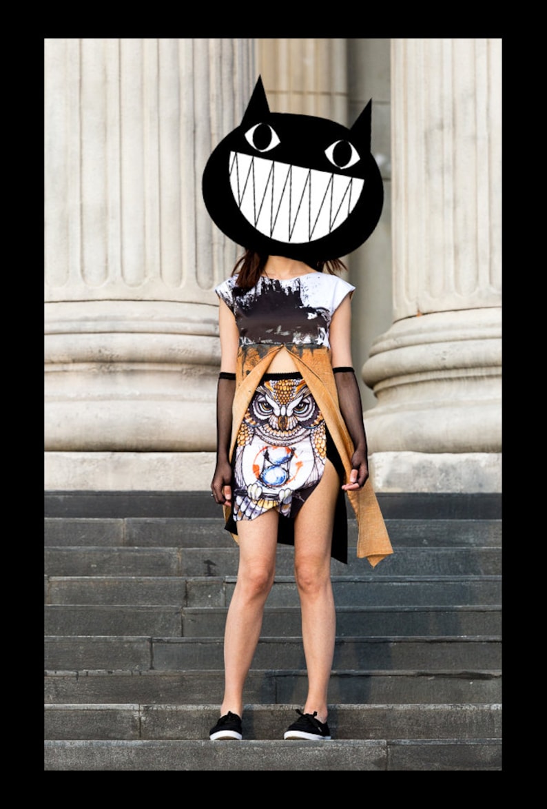 Peut inclure: Une femme portant un t-shirt graphique noir et blanc avec un masque de chat noir arborant un large sourire blanc. Elle porte une jupe marron et noire avec un motif de hibou et des gants en r&eacute;sille noirs. Elle est debout sur un escalier.