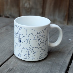 May include: White ceramic mug with a blue outline design of elephants. The elephants are clustered together and appear to be playing.