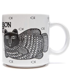 May include: White ceramic mug featuring a black and white illustration of a cat with fish-like fins and tail. The mug has the text "Larsson Cat" and small fish icons. The mug has a white handle.