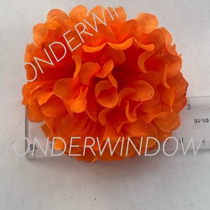 Op de afbeelding: Fel oranje kunstbloem met gelaagde bloemblaadjes. Een liniaal ligt naast de bloem, met de tekst "YONDERWINDOW" op de liniaal. De bloem is een opvallend decoratief item.
