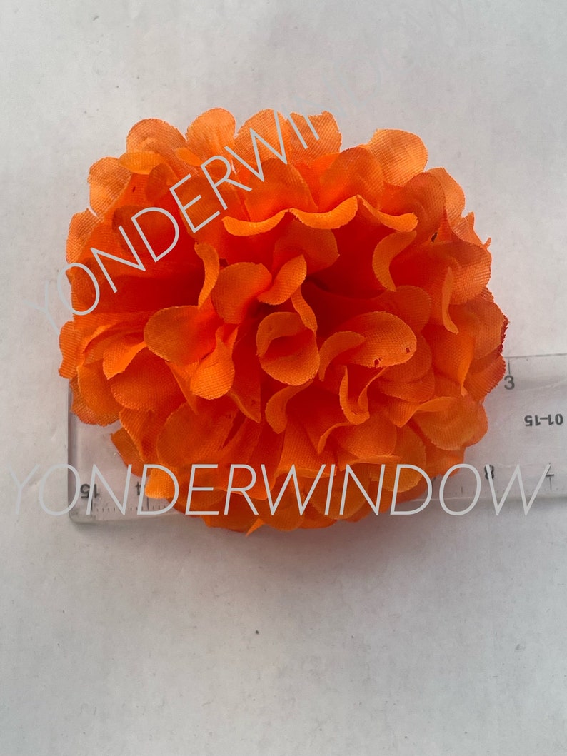 Op de afbeelding: Fel oranje kunstbloem met gelaagde bloemblaadjes. Een liniaal ligt naast de bloem, met de tekst "YONDERWINDOW" op de liniaal. De bloem is een opvallend decoratief item.
