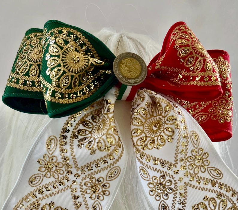 May include: A hair bow featuring green, white, and red ribbon sections with gold glitter floral patterns. The center is adorned with a circular metallic coin-like emblem featuring an eagle design. The bow has two long, dangling ribbon tails.