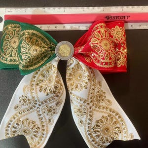May include: A decorative hair bow featuring green, white, and red ribbon sections with gold glitter floral patterns. A circular silver-toned coin is centered on the bow. A Westcott ruler placed above shows the bow is approximately 7 inches wide.