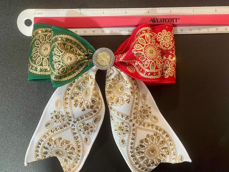 May include: A decorative hair bow featuring green, white, and red ribbon sections with gold glitter floral patterns. A circular silver-toned coin is centered on the bow. A Westcott ruler placed above shows the bow is approximately 7 inches wide.