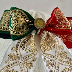 May include: A hair bow featuring green, white, and red ribbon sections with gold glitter floral patterns. The center is adorned with a circular metallic coin-like emblem featuring an eagle design. The bow has two long, dangling ribbon tails.