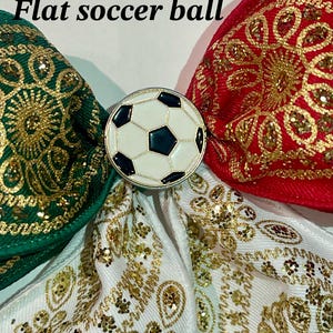 May include: A hair bow featuring green, red, and white fabric ribbons with gold glitter patterns. A circular metal charm with a black and white soccer ball design is centered on the bow. The text Option 3 Flat soccer ball is visible.