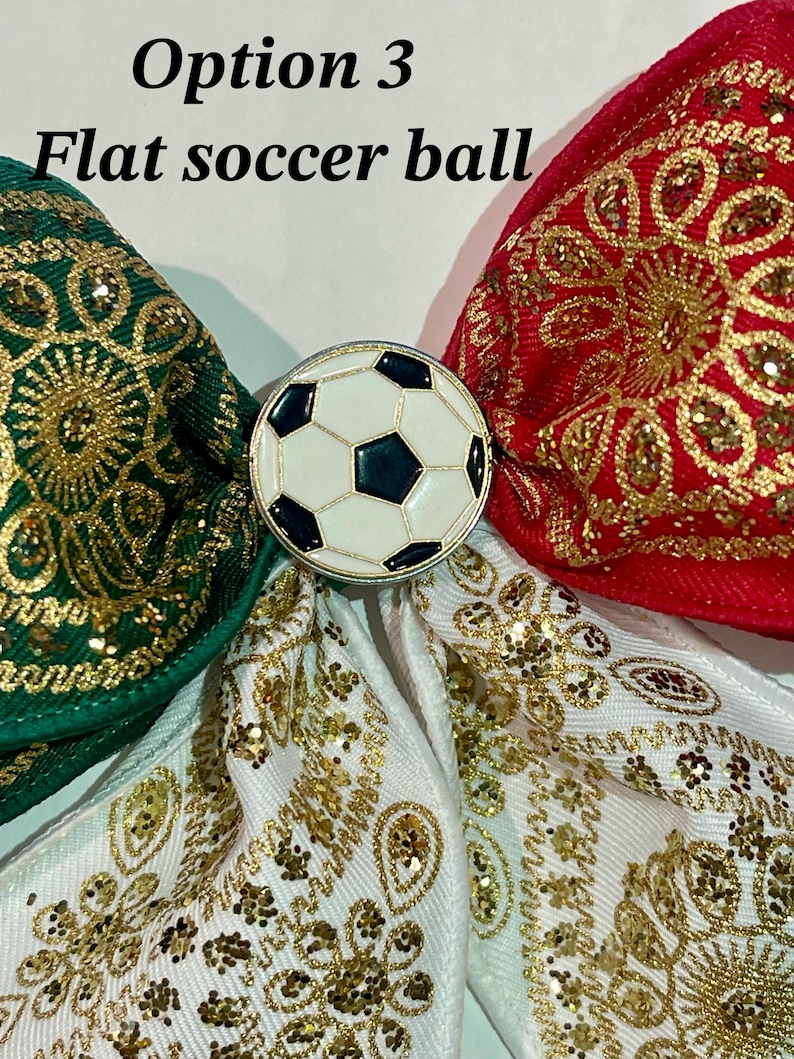 May include: A hair bow featuring green, red, and white fabric ribbons with gold glitter patterns. A circular metal charm with a black and white soccer ball design is centered on the bow. The text Option 3 Flat soccer ball is visible.