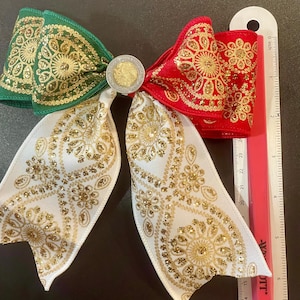 May include: A decorative hair bow featuring green, red, and white ribbon with gold glitter floral patterns. A circular coin is centered at the knot. A ruler placed alongside shows the bow measures approximately 6 inches in total width.