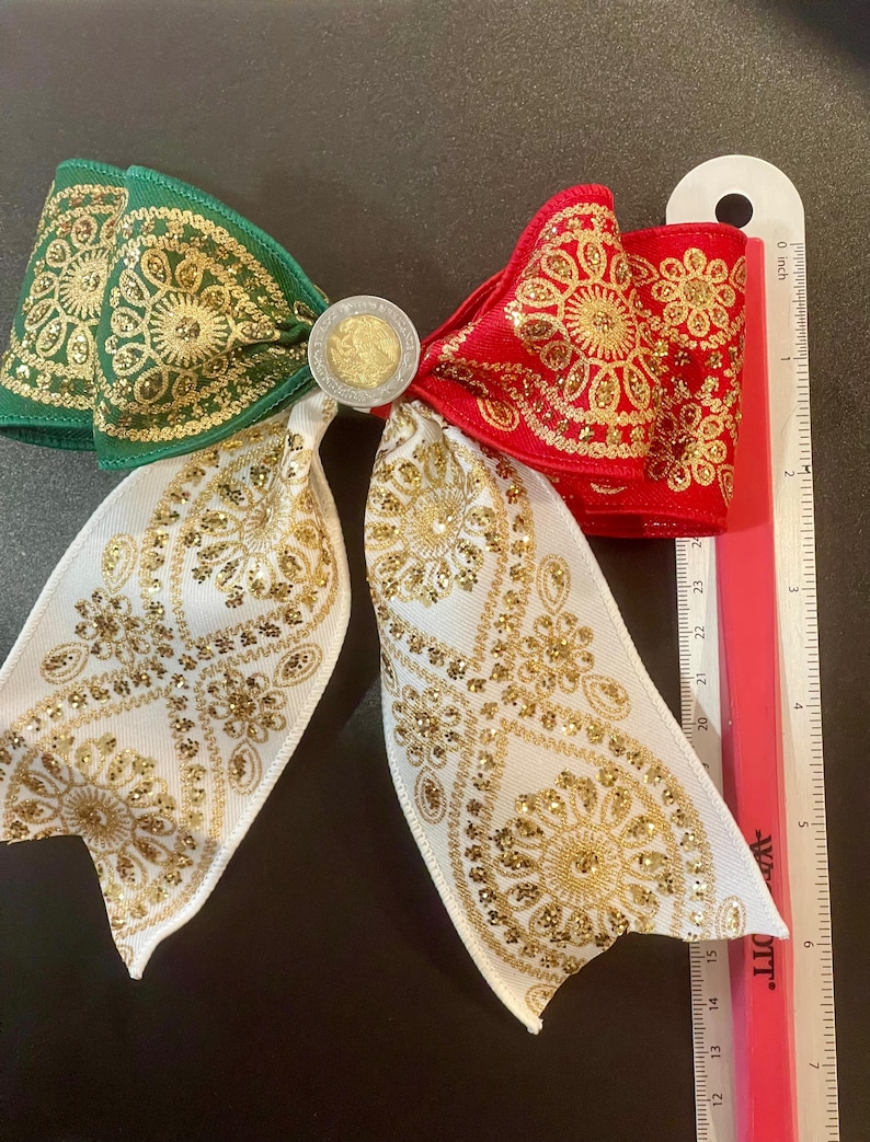 May include: A decorative hair bow featuring green, red, and white ribbon with gold glitter floral patterns. A circular coin is centered at the knot. A ruler placed alongside shows the bow measures approximately 6 inches in total width.