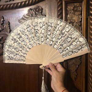 May include: A white lace hand fan with a wooden handle and a white tassel. The fan is closed and has a delicate lace pattern.