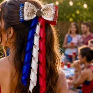 May include: A patriotic hair bow featuring red, white, and blue ribbons. The bow has a decorative center and is attached to the hair. The ribbons are curled and hang down, creating a festive look. The bow is worn by a person at an outdoor gathering.