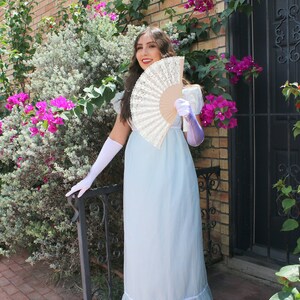 May include: A woman in a light blue dress with a white lace trim holds a white lace fan in front of her. She is wearing white gloves and purple wrist cuffs. The background is a brick wall with a green vine and pink flowers.