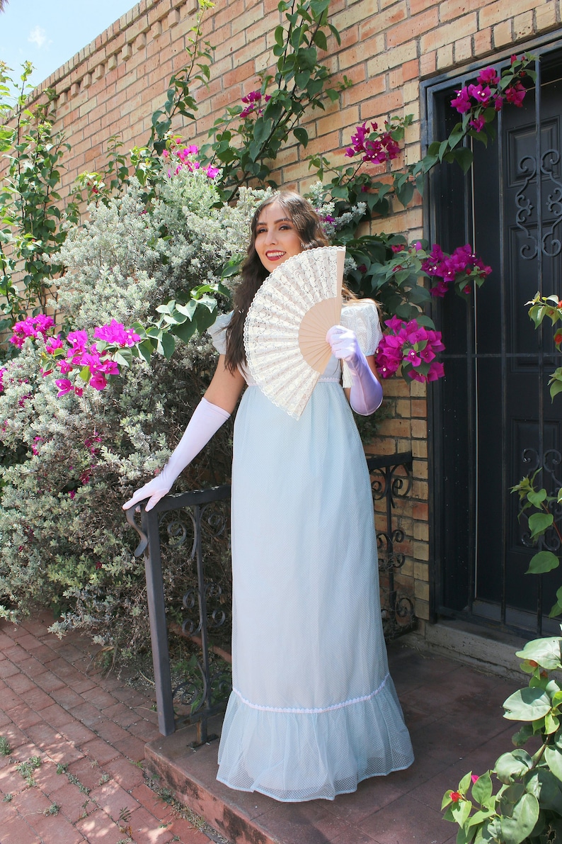 May include: A woman in a light blue dress with a white lace trim holds a white lace fan in front of her. She is wearing white gloves and purple wrist cuffs. The background is a brick wall with a green vine and pink flowers.
