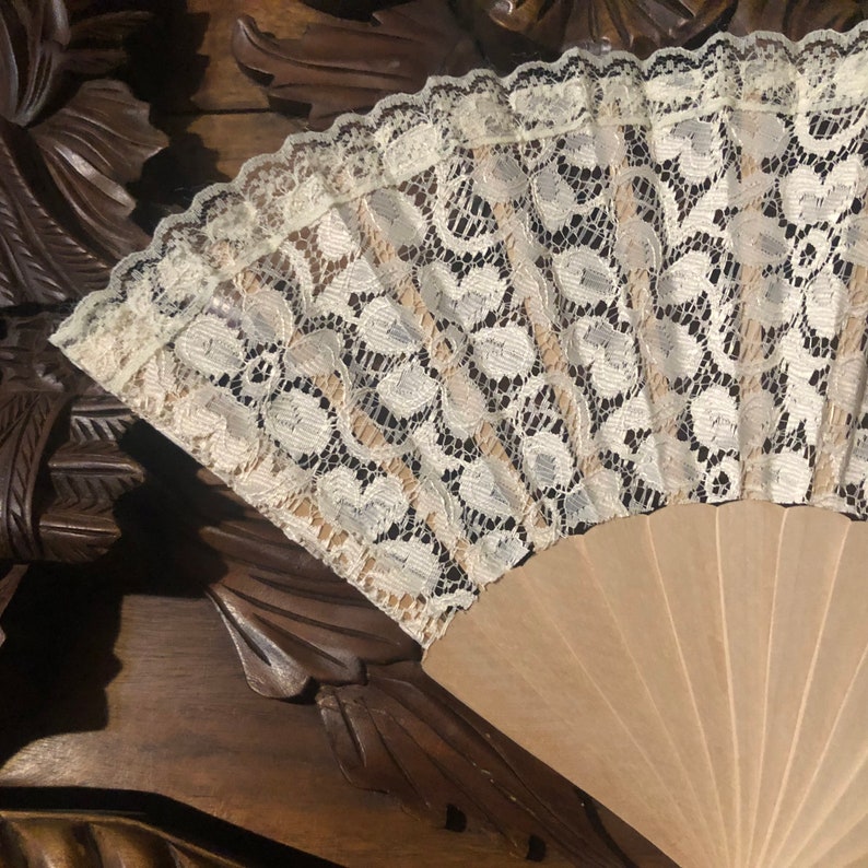 May include: A white lace fan with a wooden handle. The fan is closed and the lace is visible. The fan is on a wooden surface.