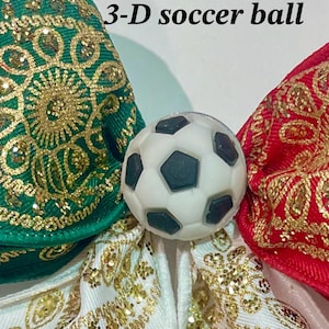 May include: A 3-D soccer ball charm centered between three fabric bows. The bows are green, red, and white, each featuring an intricate gold glitter pattern. The text 'Option 2 3-D soccer ball' is printed in black above the soccer ball charm.