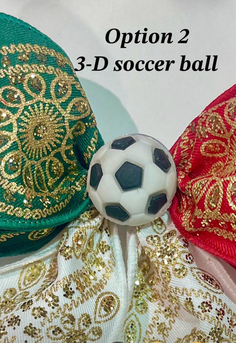 May include: A 3-D soccer ball charm centered between three fabric bows. The bows are green, red, and white, each featuring an intricate gold glitter pattern. The text 'Option 2 3-D soccer ball' is printed in black above the soccer ball charm.