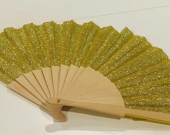 May include: A handheld folding fan featuring a bright yellow glitter fabric leaf attached to a light-colored wooden frame. The fan is fully extended in a semi-circular shape, showcasing the textured, sparkling surface of the material and the ribbed wooden structure at the base.