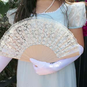 May include: A white lace fan with a wooden handle. The fan is held by a person wearing a light blue dress and white gloves.