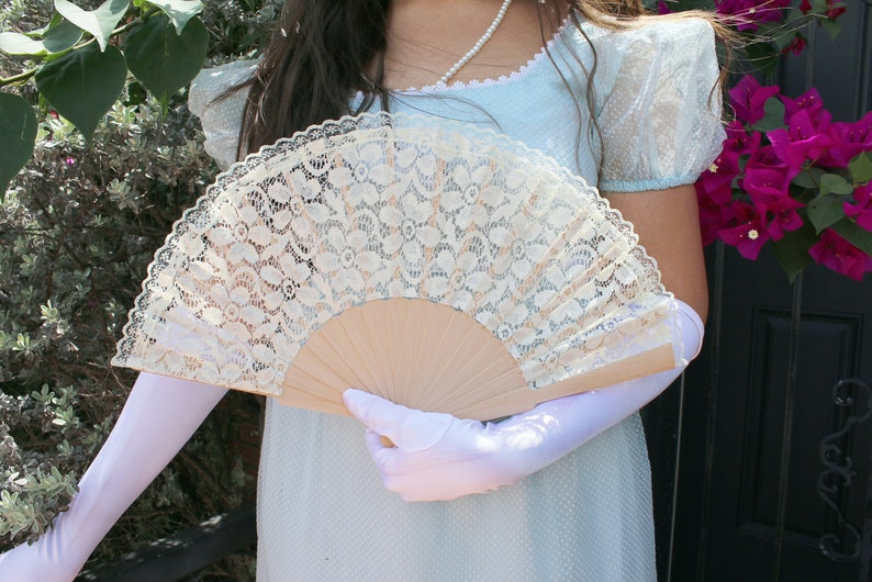 May include: A white lace fan with a wooden handle. The fan is held by a person wearing a light blue dress and white gloves.