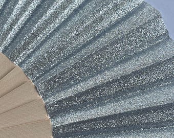 May include: A handheld folding fan featuring a pleated silver glitter surface that catches the light. The base of the fan is constructed from light beige wooden slats that radiate outward to support the shimmering, textured silver fabric material.