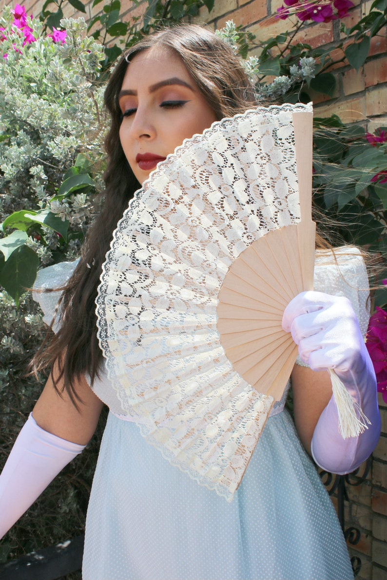 May include: A woman in a light blue dress holds a white lace fan with a wooden handle. She is wearing white gloves and has long brown hair.