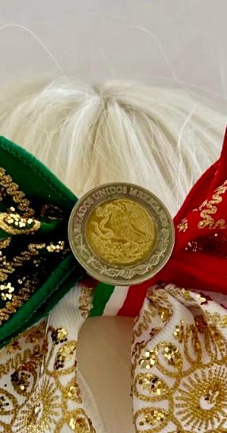 May include: A hair bow featuring green and red velvet fabric with gold glitter accents, centered with a Mexican coin displaying the text ESTADOS UNIDOS MEXICANOS and an eagle emblem. The bow includes white ribbon with gold floral patterns.