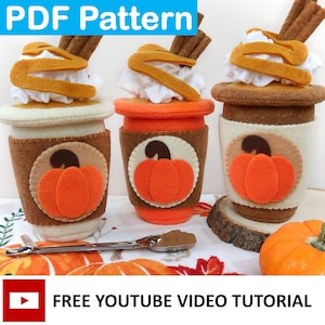 May include: Three felt pumpkin spice latte cups with whipped cream and cinnamon sticks on top. The cups have a brown felt base, a white felt middle, and a brown felt top with a pumpkin design. There is a small spoon with cinnamon on a white and orange patterned towel. The text "FREE YOUTUBE VIDEO TUTORIAL" is at the bottom of the image.