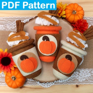 May include: Three felt pumpkin spice latte cups with whipped cream and cinnamon sticks. The cups are decorated with a pumpkin design and are sitting on a wooden surface.