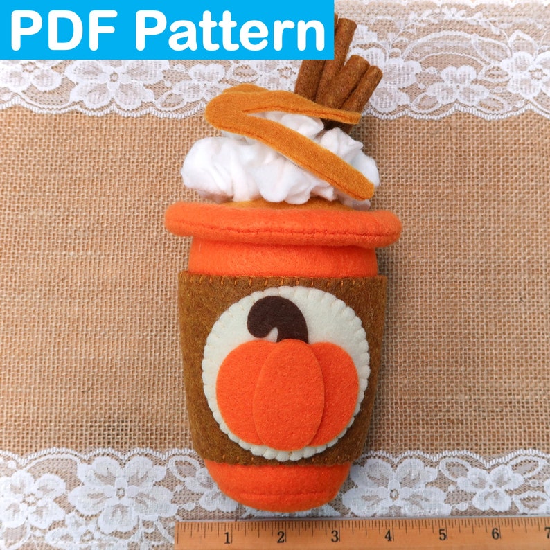May include: A felt pumpkin spice latte with whipped cream and cinnamon sticks. The latte is in a brown paper cup with a pumpkin design. The cup is sitting on a burlap background.