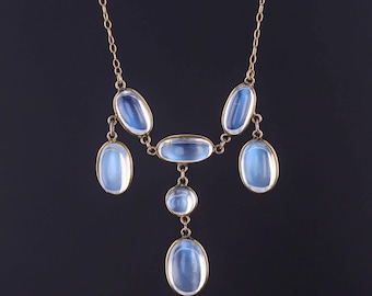 Könnte beinhalten: Eine goldfarbene Halskette mit fünf ovalen, durchscheinenden, hellblauen Cabochon-Steinen in einem Tropfenmuster. Die Steine sind in Metallfassungen eingefasst und durch zarte goldfarbene Kettenglieder verbunden, was ein symmetrisches Anhängerdesign ergibt.
