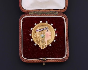 May include: A gold brooch featuring a profile of a woman with flowing hair, adorned with a jeweled headband and earrings. The brooch is surrounded by small pearls and set in a red velvet-lined box. The woman's face is painted with delicate features.