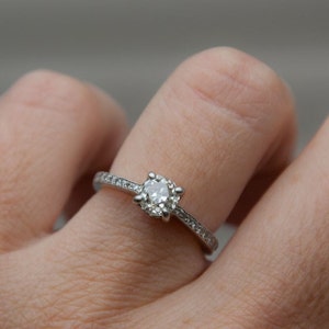 Puede incluir: Un anillo de compromiso de plata con un solo diamante redondo engastado en garras. La banda es fina y tiene peque&ntilde;os diamantes engastados en pav&eacute;.