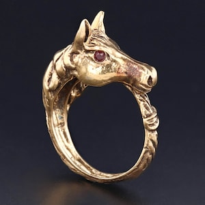 Puede incluir: Un anillo de tono dorado con un diseño detallado de cabeza de caballo. El ojo del caballo está engastado con una piedra preciosa roja.