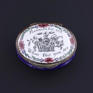 May include: A small, oval-shaped, blue and white enamel trinket box with a silver lid. The lid features a black and white illustration of a basket of flowers with the text "Remember me when this you see".