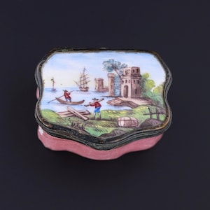May include: A small, pink, rectangular box with a hinged lid. The lid is decorated with a hand-painted scene of a boat with two figures in it, sailing past a fortified structure on a body of water. The scene is surrounded by a black border.