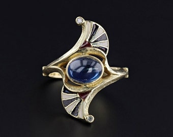 Puede incluir: Un anillo de oro con una piedra preciosa azul y detalles de esmalte blanco y rojo. El anillo está diseñado en una forma geométrica estilizada.