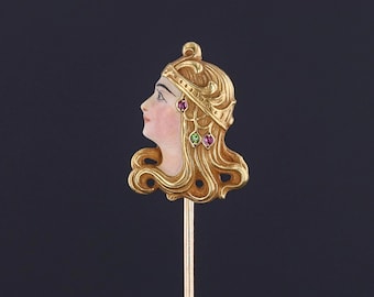 May include: A gold-toned stick pin with a detailed enamel portrait of a woman wearing a crown and earrings. The woman's hair is styled in a wavy, Art Deco design.
