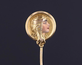 May include: A gold-toned stick pin with a round, raised design featuring a woman's profile. The woman is wearing a crown and has long, flowing hair.