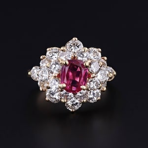 May include: A gold ring featuring a large, oval-cut ruby center stone surrounded by a halo of round, brilliant-cut diamonds. The ring's design is set against a black background, highlighting the sparkle of the gemstones.