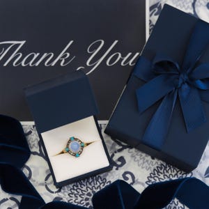 Puede incluir: Una caja de anillo azul con un interior blanco y un anillo con una piedra azul y acentos turquesa. La caja est&aacute; sobre una superficie estampada con una cinta azul y una tarjeta que dice "Gracias".