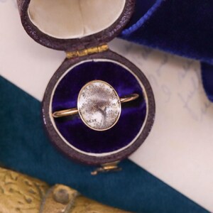 K&ouml;nnte beinhalten: Ein goldener Ring mit einem wei&szlig;en ovalen Stein in einer Ringbox. Der Stein hat ein graues und wei&szlig;es Muster.