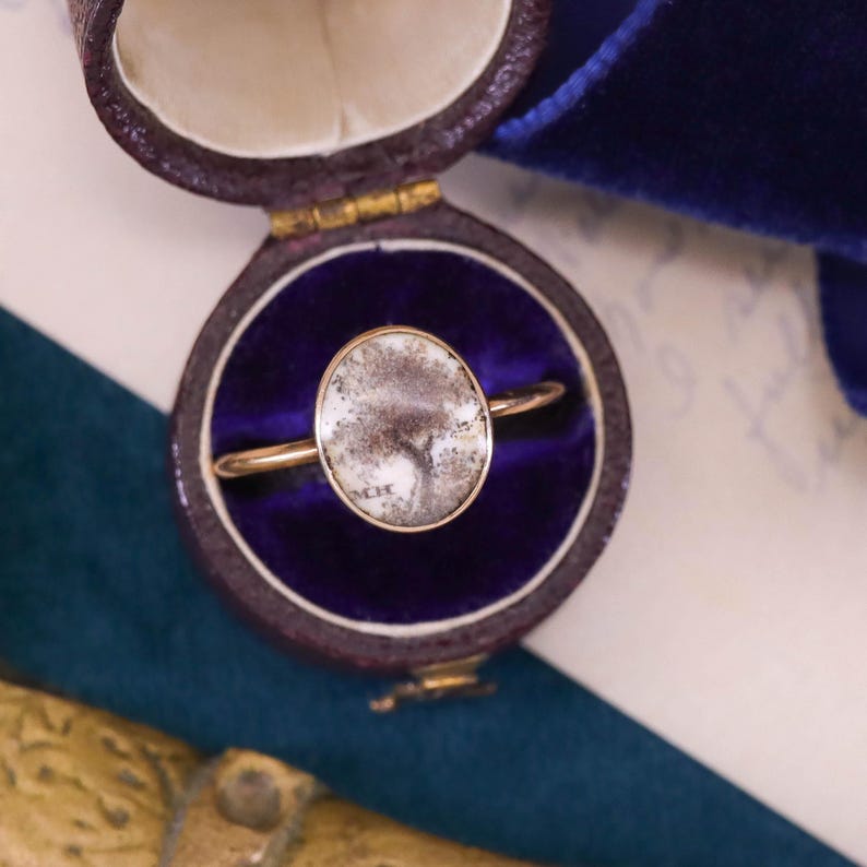 K&ouml;nnte beinhalten: Ein goldener Ring mit einem wei&szlig;en ovalen Stein in einer Ringbox. Der Stein hat ein graues und wei&szlig;es Muster.