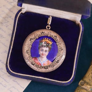 Pu&ograve; includere: Un ciondolo a medaglione d'oro con uno sfondo in smalto blu e un ritratto dipinto di una donna che indossa una corona di fiori. Il medaglione &egrave; decorato con intricati motivi floreali.