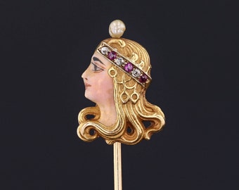 May include: A gold-toned stick pin with a detailed enamel portrait of a woman with a crown of pink and white gemstones. The pin is on a black background.