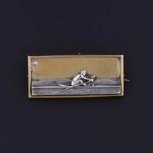 May include: A rectangular gold-tone brooch with a silver-tone skier figure. The brooch features a small diamond and a two-tone design, with a gold upper section and a silver lower section depicting a snowy landscape.